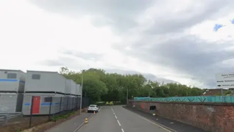 Google General view of Foundry Lane in Smethwick. On the left-hand side there are industrial units and on the right-hand side there is a wall. In the background there is a row of trees