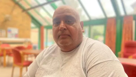 Simon Thake/BBC A man with a shaved head wearing a grey t-shirt sits in a room with a glass roof and wall and red chairs at tables behind him