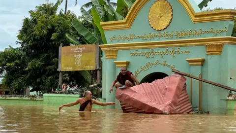 Getty Images A Buddhist monk wades through floodwaters as another sits on a damaged roof in front of a monastery in Sin Thay village in Pyinmana, Naypyidaw region of Myanmar, on September 13, 2024, following heavy rains following Cyclone Yagi.