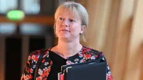 Shona Robison, who has blonde hair, looks to her right while walking towards the camera. She is carrying a black folder held against her chest. She is wearing a red and black floral top. 