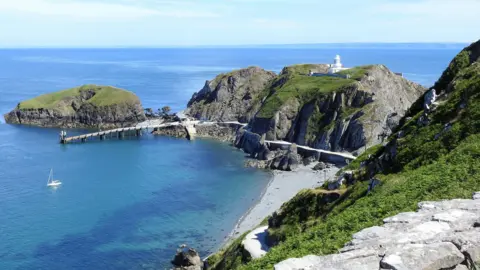 Getty Images Looking down on the landing stage where the ferry docks from the path up to the top of Lundy island. The South lighthouse is on a large rock on the right and there is a white yacht moored in the harbour in the clear blue sea.