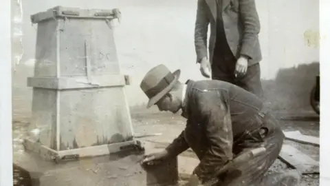 Ordnance Survey A black and white image of a man wearing a bowler hat skimming the concrete off a trig pillar being built. Another man is standing next to him holding a cigarette.