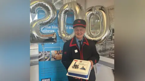 Southeastern An image of Mark Jones during the Railway 200 celebrations. Mark is wearing a black and red train dispatcher uniform, and is holding a cake. He is stood in front of three balloons spelling out the number 200. 