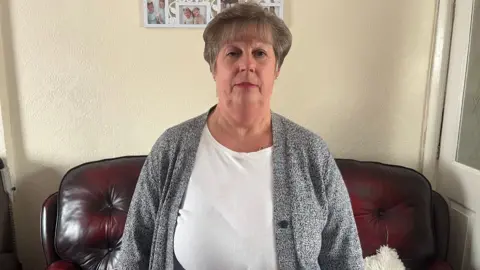 Jean Cross in her living room, with a dark brown sofa behind her and photos of family on the wall