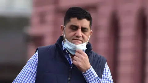 Spindrift A dark haired man in a check shirt and gilet with a facemask pulled down on his chin