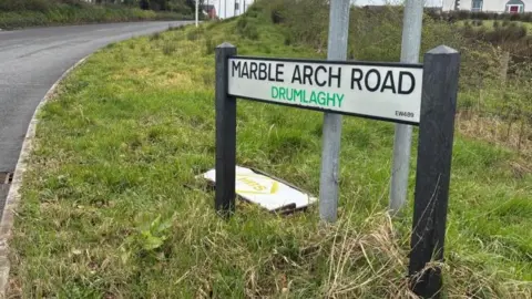 A road sign in grass. It says Marble Arch Road in block capital letters.