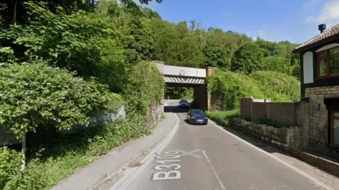 Google A Google Maps screengrab of Winsley Hill showing two cars, trees, a cottage and a road bridge.