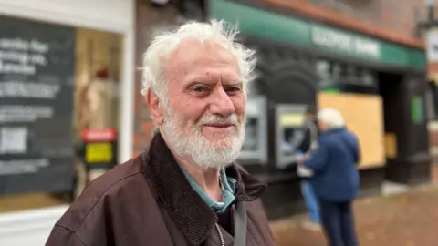 BBC / George Carden An old man with white hair. He is wearing a dark brown jacket and a blue shirt. He is standing near a bank.