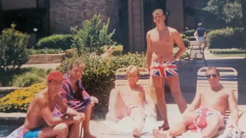 Gary Allen A photograph of five men around a pool in their swimming trunks. One of the men stands up wearing Union Jack swimming trunks.