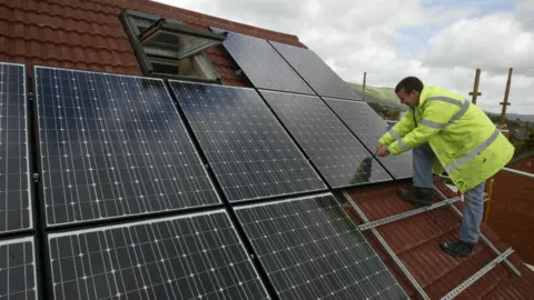 PA Media A man wearing a hi-vis jacket fixing dark solar panels to a slate roof.
