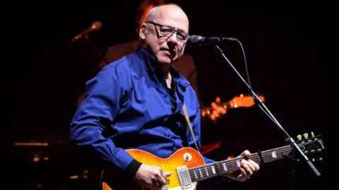 Mark Knopfler performing onstage. He is a bald man with glasses, wearing a blue shirt, and playing a guitar. A microphone is in front of him.