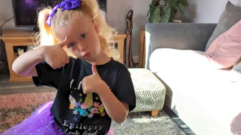 Noa-Rose posing with her thumbs up in her living room. She wears a black 'Epilepsy looks good' t-shirt, a purple sparkly skirt and has her hair in bunches with purple bows.