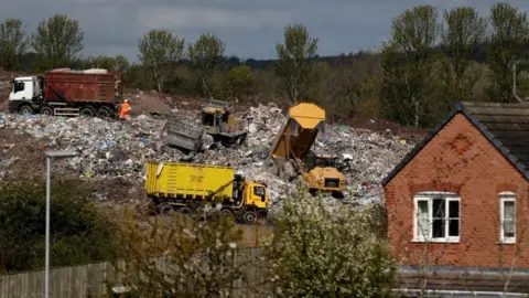 A landfill site with lorries and waste trucks driving over it. On the other side of a nearby fence is a house.