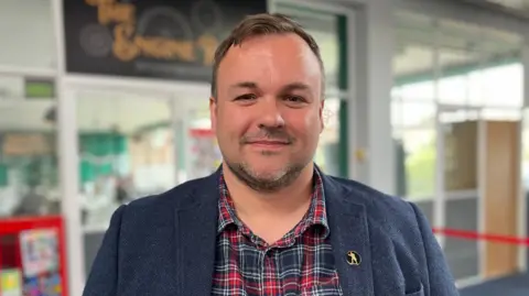 Shaun Whitmore/BBC Terry Jermy with short, brown hair, smiles for the camera. He is wearing a red and black plaid shirt under a grey jacket, with the windows and the sign of a shop seen behind him in soft focus.