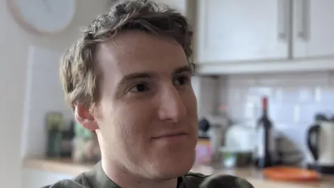 A man is in a kitchen. He has light-coloured, reddish hair and is looking off to the right with a wry, slight smile on his face. He is wearing a green-coloured t-shirt. In the background, white kitchen cupboards and appliances can be seen