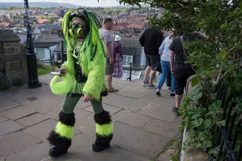 Getty Images A costumed woman walking on a stone-paved walkway overlooking a town with rows of red-tiled roofs and closely packed buildings. In the distance, low hills and greenery are visible beyond the town. The woman dominating the foreground is wearing a fluorescent green and black outfit with a futuristic, industrial aesthetic. Her costume includes a gas-mask-style face covering with round filter elements. She has green synthetic hair extensions, a green-and-black fur-textured jacket, a green mesh garment visible at the torso and legs.