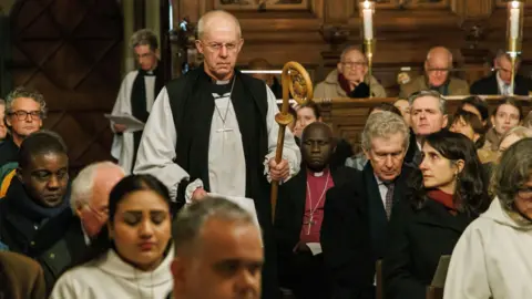 Lambeth Palace/PA Justin Welby, arrives for a service of Evensong at Lambeth Palace Chapel. He carries the crozier and walks toward the altar, with the pews full of congregants on both sides of the aisle.