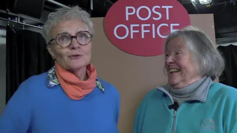 Jo wears a blue jumper and pink shawl. She wears black rimmed round glasses and has short grey hair. Pam wears a turquoise fleece with a grey polo neck underneath. She has grey hair in a bob and is smiling at Jo as she speaks.