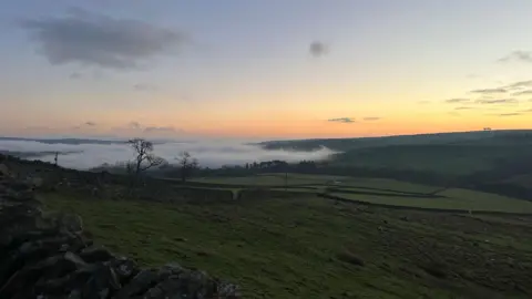 BBC Weather Watchers/Iloveweardale A layer of fog over a valley of fields. The sky above the horizon is orange, turning into light blue. There are a few clouds in the sky and the ground is dark.