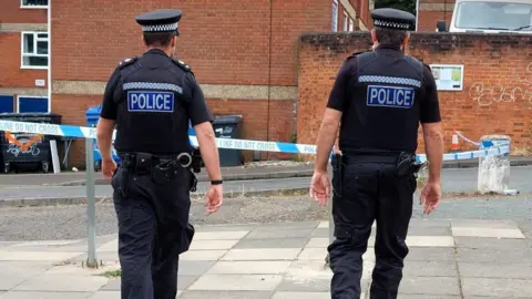 Norfolk Police Two police officers dressed in black with their backs turned to the camera as they walk. They are walking on concrete paving slabs, and in the background, is a road, some bins, and brick walls and apartment blocks.