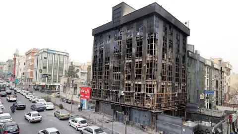 Getty Images Cars pass by a burnt building destroyed during public protests in the Iranian capital Tehran. There are lots of cars and some two-wheelers on the road. 