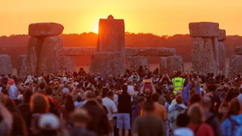 English Heritage Thousands of people gathered in a crowd - many holding up cameras and phones - watching the sun rise up behind the stones at Stonehenge, with the silhouettes of trees and a yellow sky in the background. 