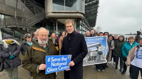 Roger Clubley and Andy Gollifer hold a Save the National Glass Centre sign in front of a group of about 20 campaigners outside the venue. Clubley has glasses, white hair and beard and is wearing a khaki coat over a green jumper. 