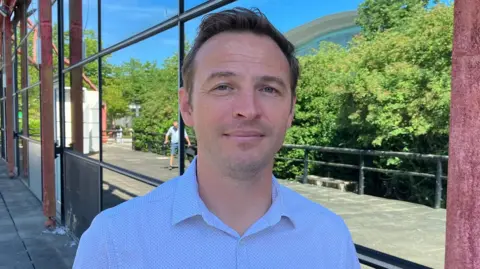 Sam Read/BBC Ryan Gillies is looking directly at the camera and smiling. He has dark blonde hair and is wearing a white patterned shirt. He is standing in front of The Point, a former cinema which closed in 2015. The outside of the building is made from a red metal structure in a pyramid shape with mirrored windows which in this picture show the reflection of a path and green trees and shrubbery. 
