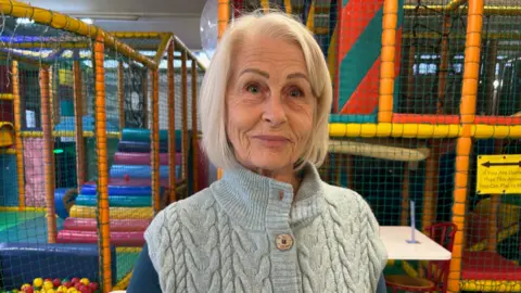 An older woman with white hair and wearing a blue cardigan stands in a child's soft play center.