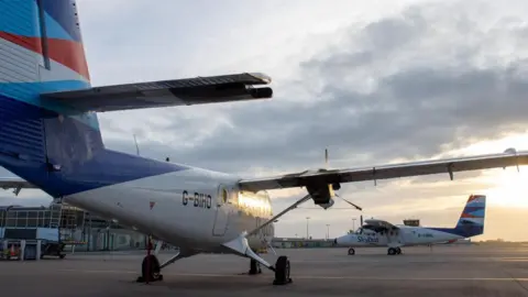 Two twin otter planes. One facing away to the camera and one facing diagonally. The two planes are on airport tarmac. The sky is grey and cloudy.
