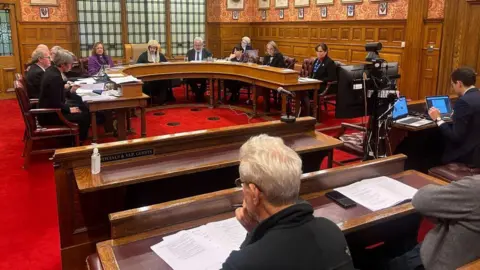 The Legislative Council chamber, which has people sitting on brown benches watching the proceedings. The MLCs, both male and female wearing smart clothes, are sitting around a brown horse-shoe shaped table.  The President of Tynwald sits in the middle wearing a wig and the Bishop sits next to him wearing purple.