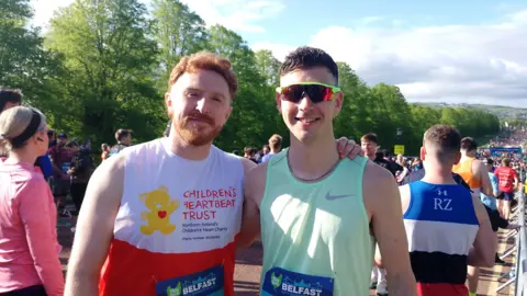 Mr Tolan, who is on the right, stands next to another man. He has short black hair and is wearing a mint green vest with glasses. To the left is a man with ginger hair and facial hair wearing a red and white running vest with 'Children's Heartbeat Trust' written on it.