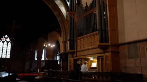 Organ custodian Stephen Lomas playing an organ in a church. The organ has dark wood panelling and the church is barely lit.