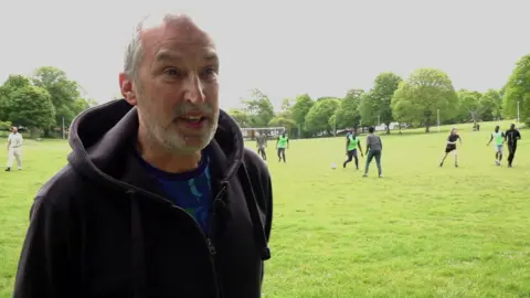 A man in a hoodie looks to the side of the frame while a game of football takes place in the grassy park behind him.