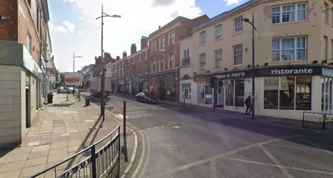 A city-centre street, lined by shops and restaurants, with lorries and cars driving down it and signage by the side.