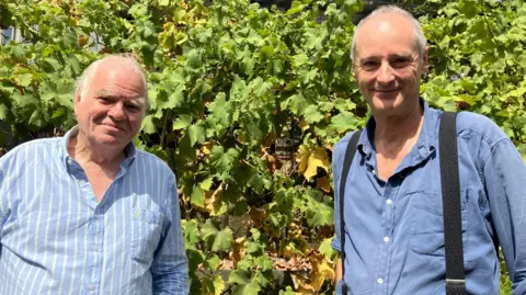 This photograph shows two men standing outdoors in front of a dense row of grapevines. In the foreground, the two men, Tony Coleman and Nick Mair are positioned side by side. Tony Coleman is on the left wearing a light blue shirt with thin vertical white stripes. Nick Mair stands on the right wearing a darker blue button-up shirt with black braces. Both are facing the camera. Behind them is a thick wall of grapevine foliage. The leaves are mostly bright green, with some turning yellow and brown. Among the leaves, small clusters of Bacchus grapes can be seen hanging from the vines.