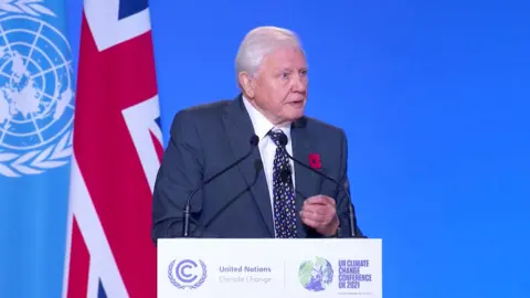 Attenborough in a suit, speaking at a United Nations climate conference lectern, with the UK flag behind him