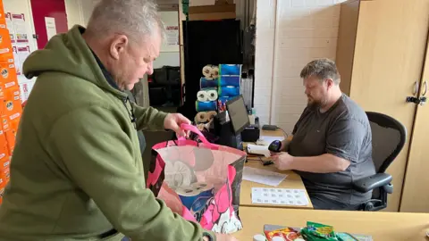 A man in a khaki hoodie putting toilet rolls in a pink carrier bag. A man in a grey T-shirt is sat at a desk scanning items. 