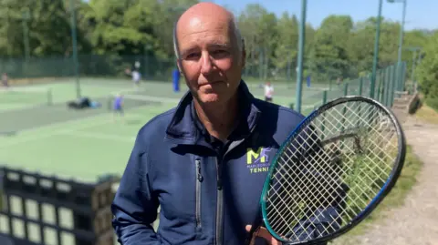 Andrew Payne - Chair, Marlborough Tennis holding his racket and wearing a Marlborough Tennis navy blue zip-up jacket with the club's tennis courts behind him