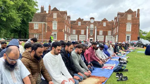 Jon Wright/BBC Rows of men kneeling on mats on the grass in front of a Tudor mansion. There is a collection of shoes on the grass in front of the mats. The Muslim men appear to be praying and some wear caps while others do not.