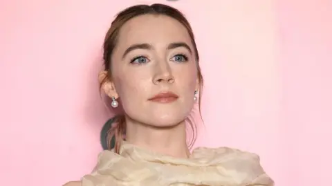 Getty Images A young woman stands in a peach coloured dress. Her hair is up and she's wearing pearl earrings. The backdrop is pink.