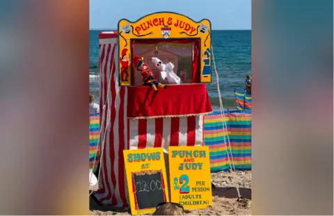 Jonathan Crump A Punch and Judy performance at the beach on a sunny day. Mr Punch and the ghost puppets are seen. There are placards underneath the performance tent, listing the showtimes and the prices of £3 per person.