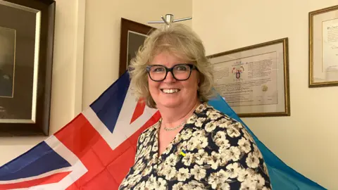 A woman with short, curly, blonde hair wears large square glasses whilst also wearing a floral patterned top. She is stood in front of a Union Flag and a Ukrainian flag. 