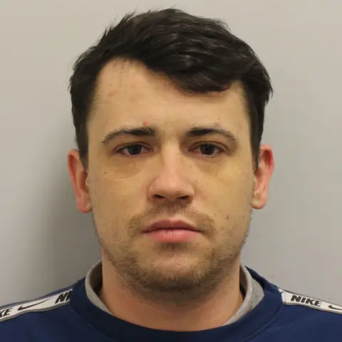 Met Police Custody image of Jordan McSweeney, a  young man with dark hair wearing a dark blue sweatshirt and grey tshirt