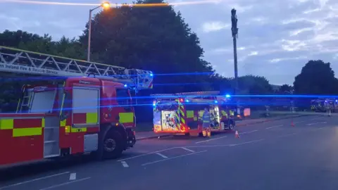 Two fire engines on a road. Fire fighters are present. A 5G mast is on fire.