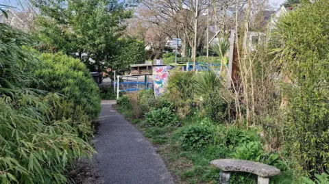 Jim Gardner A footpath leading to a road is bordered by large shrubs and a stone bench.