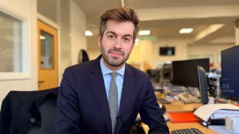BBC Correspondent Joe Pike sitting at his desk, wearing a suit and looking into the camera