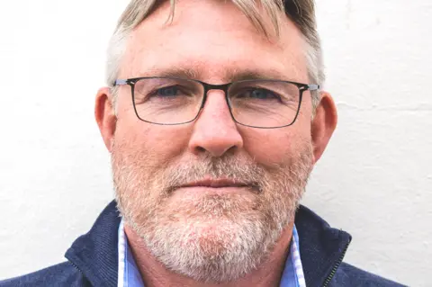 Catch at the Old Fish Market A head and shoulders shot of Sean Cooper standing against a white wall. He has short hair and a stubble beard. He is wearing black rimmed glasses, a navy blue zip-up jacket and light blue open collar shirt.