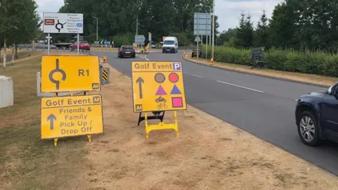BBC Three yellow signs are on the left near a road on the right. Two of them include the words golf event, with one also saying friends & family pick up/drop off and the other having a number of symbols by an arrow sign and a P parking symbol. A roundabout and trees are in the background.