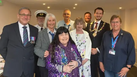 A mixed group of people stand together smiling. One man has military medals fixed to his chest, and another wears a Navy-style white uniform. One man wears a ornamental mayoral necklace and a suit.